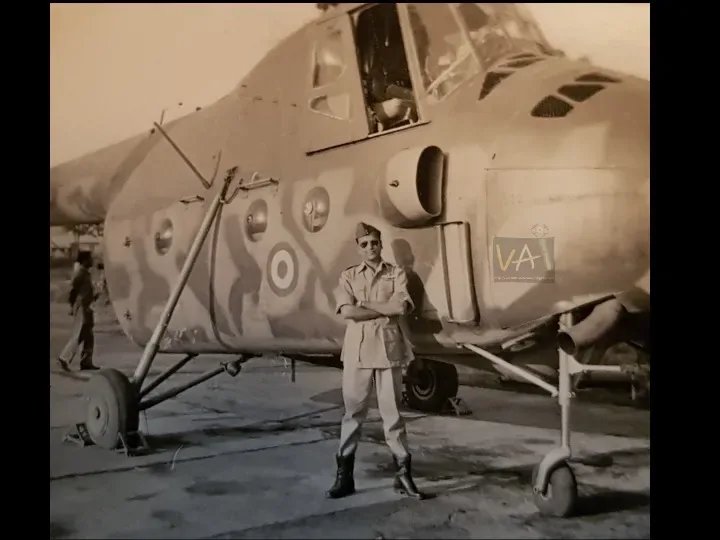 A young Gp Capt R V Singh standing with arms crossed in front of a camouflaged Mi-4 helicopter, an airman visible in the background