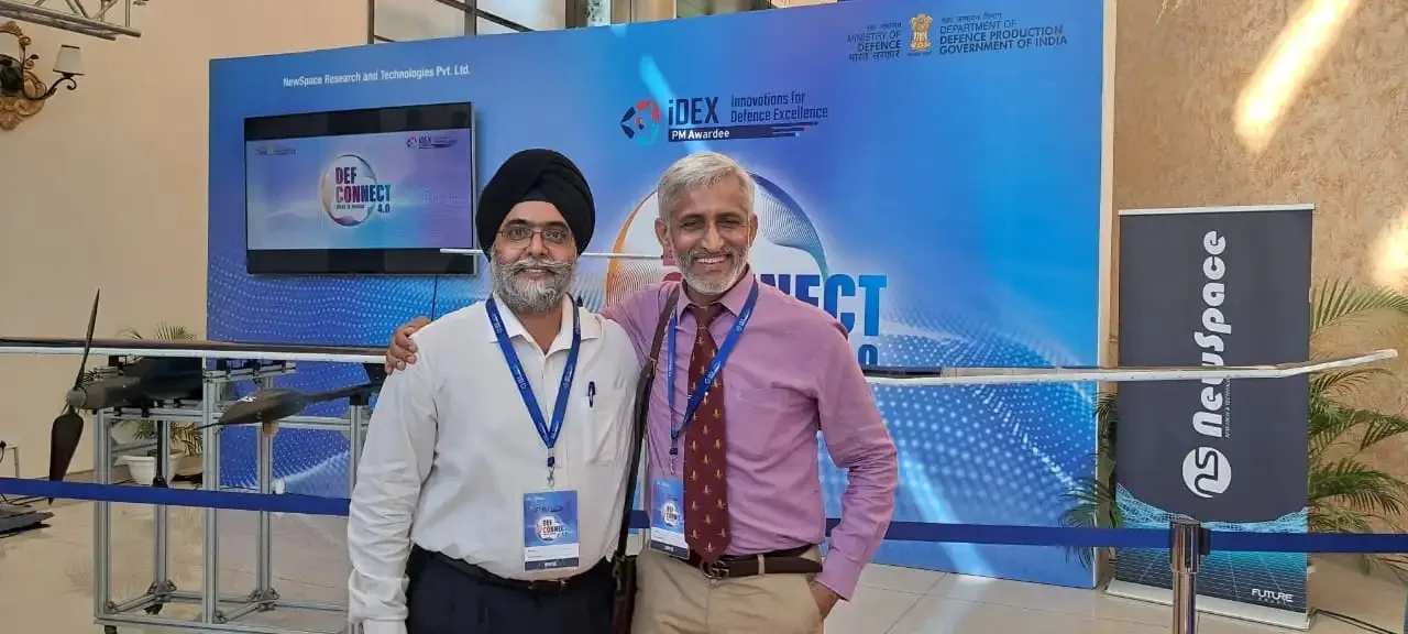 Two attendees posing together in front of the iDEX and Def Connect 4.0 backdrop, with a drone displayed nearby
